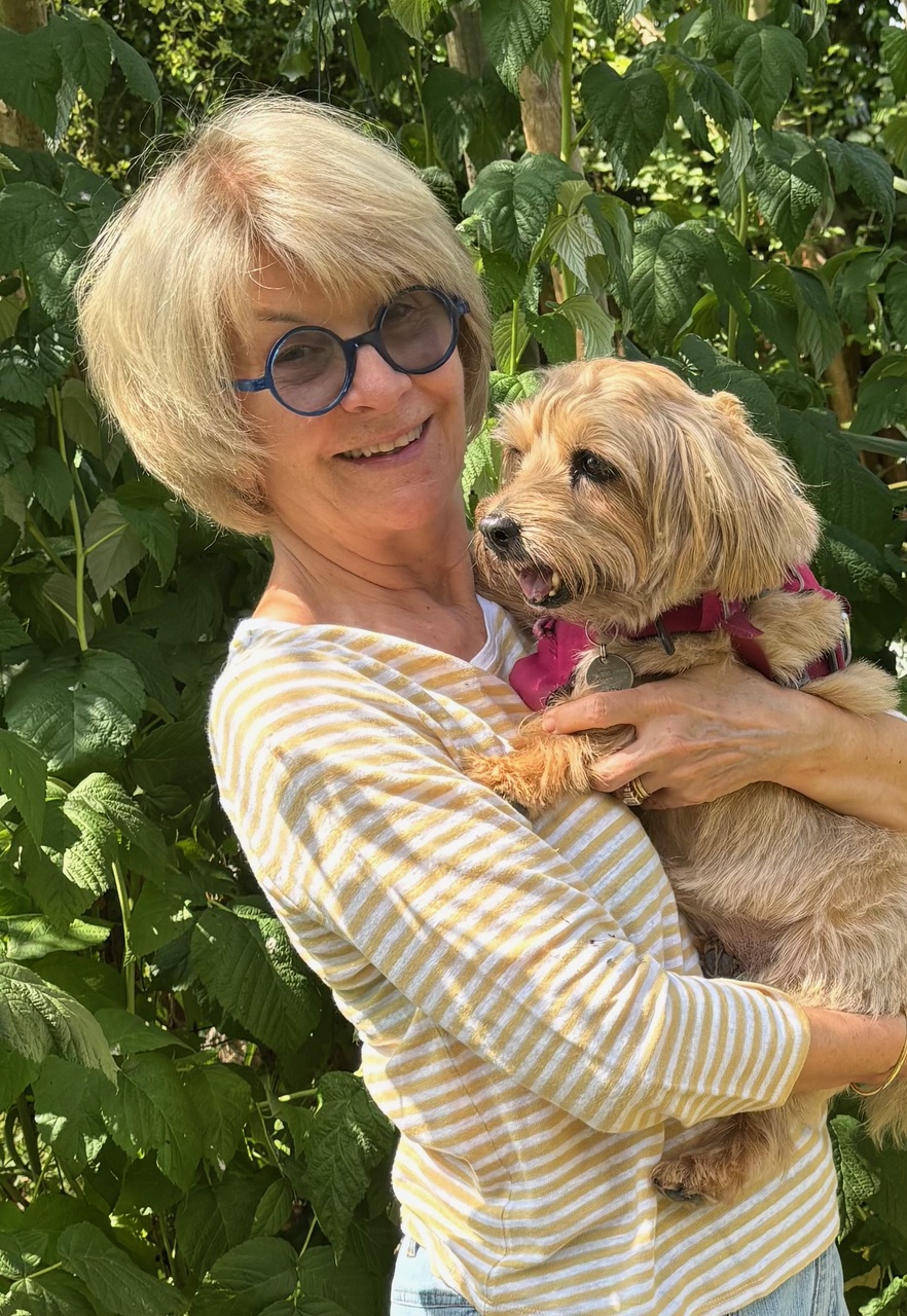 A woman with short hair and round blue glasses smiles while holding a small tan dog wearing a pink harness. They are outdoors, surrounded by lush green leaves.